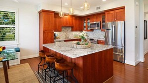 A modern kitchen with wooden cabinets, stainless steel appliances, and a central island with bar stools. - Pili Mai 12L Central AC By Suite Paradise (Koloa)