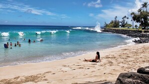 A beach with clear blue water, people swimming and surfing, and a person sitting on the sand. - Pili Mai 12L Central AC By Suite Paradise (Koloa)