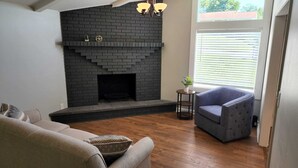 Una sala de estar con chimenea de ladrillo, un sillón azul, un sofá y una mesita con una planta.