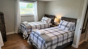 Un dormitorio con dos camas, cada una con cubrecama y almohadas a cuadros, una mesita de noche con lámpara, y una ventana con persianas.