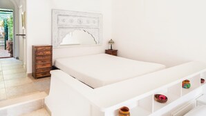 A white bed with a headboard, a small wooden nightstand with a lamp, and a shelf with decorative items.