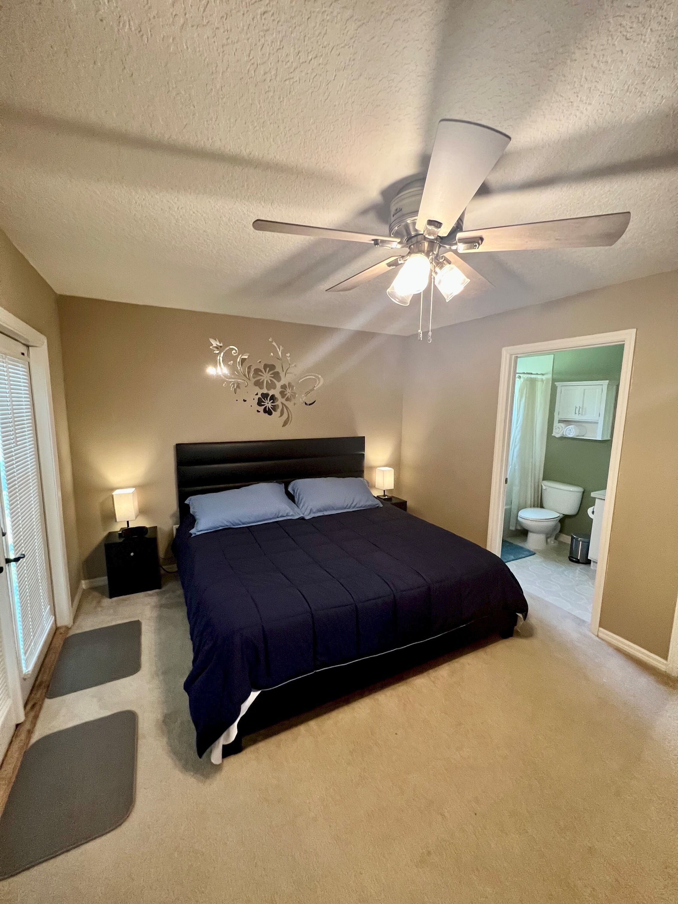 Une chambre à coucher avec un lit, des tables de chevet, un ventilateur de plafond et une salle de bain visible à travers une porte ouverte.