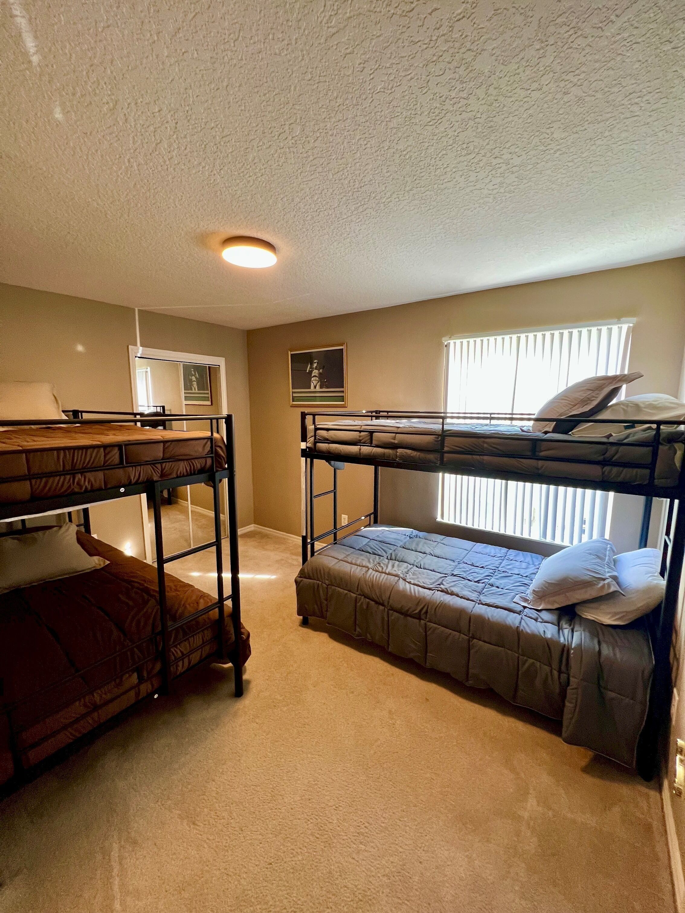 A room with two bunk beds, a small kitchenette, a framed picture on the wall, and a window with blinds.