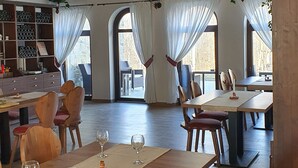 A spacious restaurant with wooden tables and chairs, large windows with curtains, and hanging plants. - Vatra Boiereasca (Cacica)