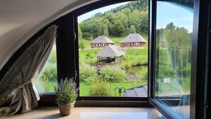 A room with a large window overlooking a green landscape with two wooden buildings and a small pond. - Vatra Boiereasca (Cacica)
