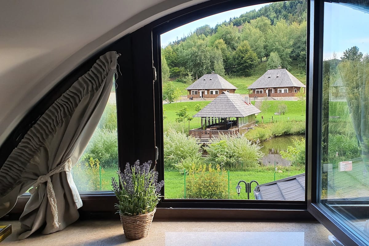 A room with a large window overlooking a green landscape with two wooden buildings and a small pond.