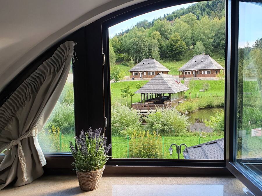A room with a large window overlooking a green landscape with two wooden buildings and a small pond.