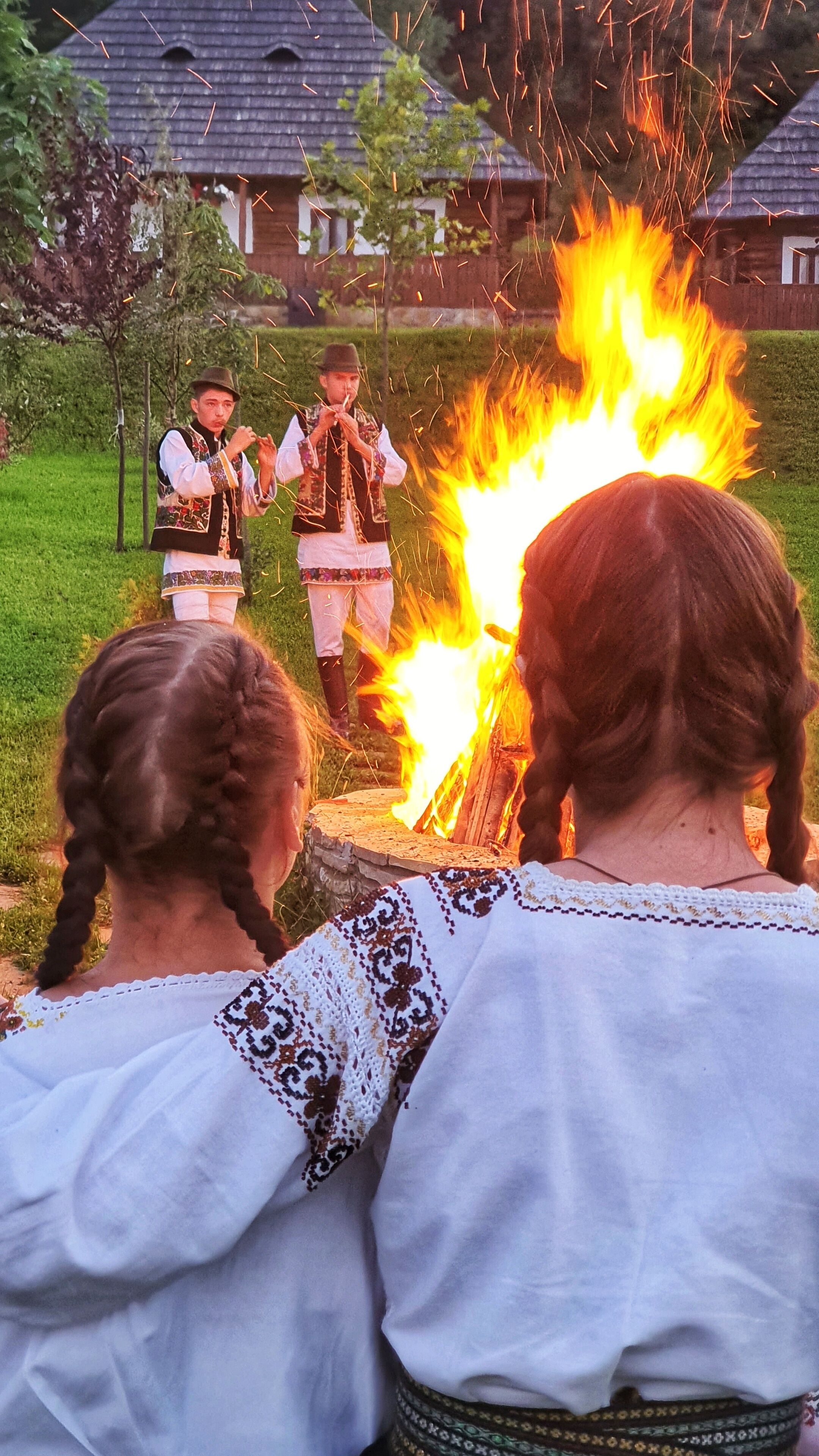Un grupo de personas vestidas con atuendos tradicionales reunidas alrededor de una fogata, con casas al fondo.