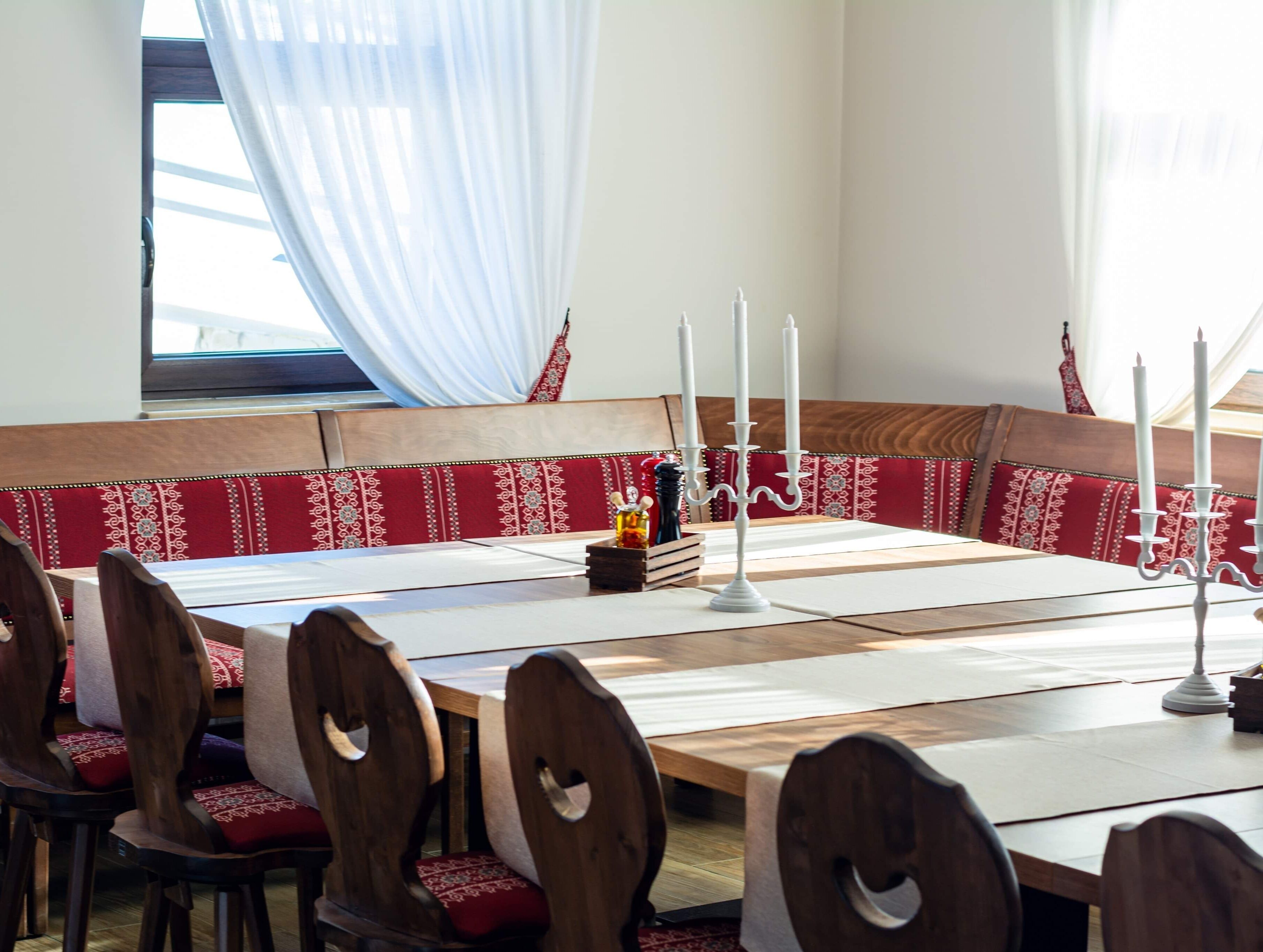 Un espace repas avec une longue table en bois, des bancs recouverts d’un tissu rouge et des bougies blanches.
