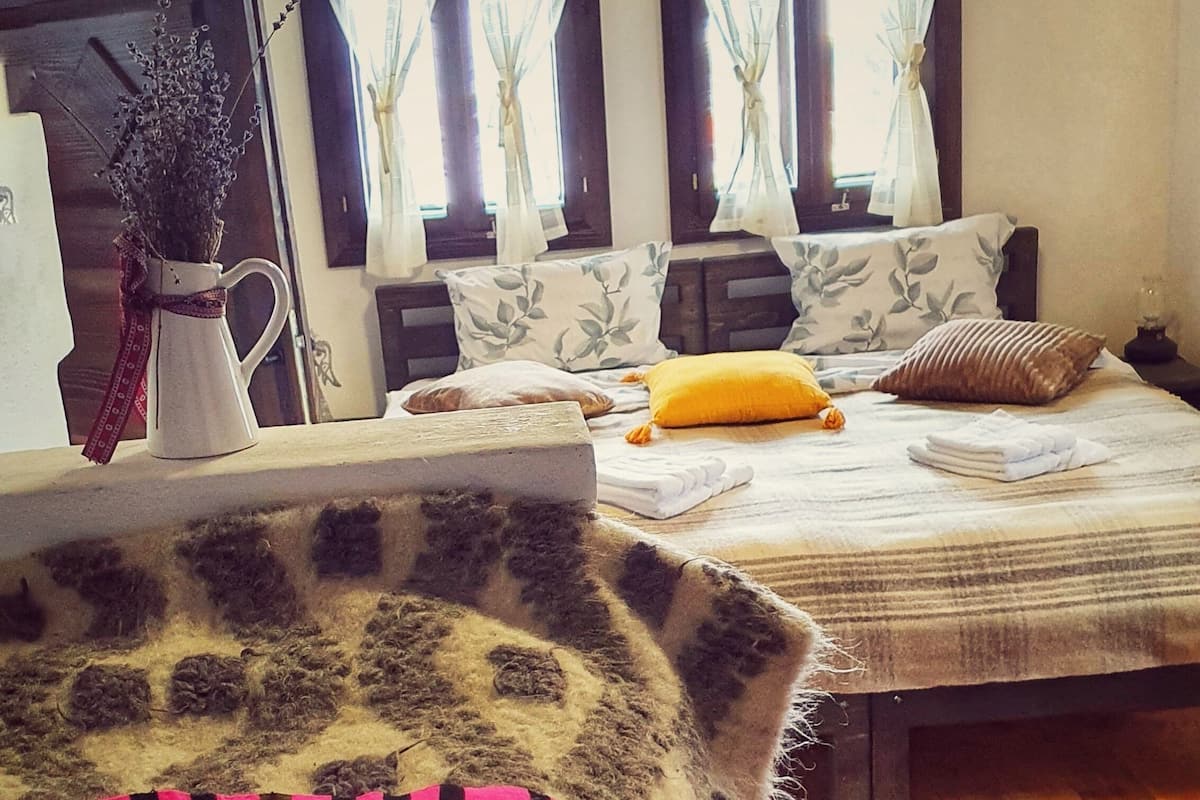 A room with a bed, a window with curtains, a vase with dried flowers, and a patterned rug.