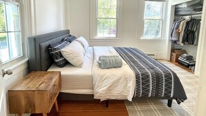 A bedroom with a bed, a wooden nightstand, a closet with hanging clothes, and two windows. - Historical 1st Floor 3BR APT Brunswick @ Bowdoin (Brunswick)
