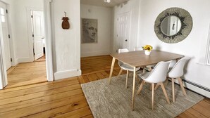 A dining area with a wooden table, white chairs, a rug, a wall-mounted mirror, and a door leading to another room. - Historical 1st Floor 3BR APT Brunswick @ Bowdoin (Brunswick)