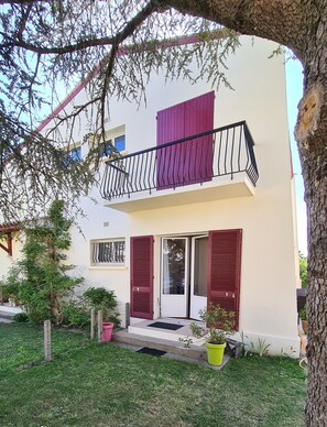 Un bâtiment à deux étages doté d’un balcon, d’une porte avec des volets et d’un petit jardin agrémenté de plantes en pot.