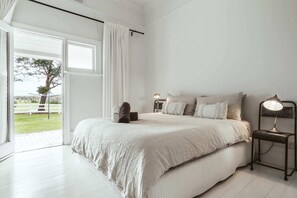 A bedroom with a large bed, a bedside table with a lamp, and a view of a grassy area outside. - 'Mayfield' - Luxury Farmhouse with Pool (Mayfield)