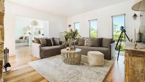 A modern living room with a sofa, round coffee table, and ottoman. There are large windows and a view of greenery outside. - 'Serenity Gerringong' Designer Beach House (Werri Beach)