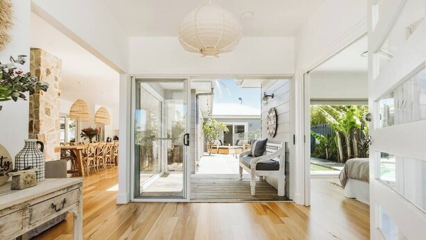 A spacious interior with a wooden floor, a dining area with a table and chairs, a sofa, and a large sliding glass door leading to an outdoor area. - 'Serenity Gerringong' Designer Beach House (Werri Beach)
