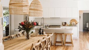 A modern kitchen with white cabinets, wooden dining table, and hanging wicker lights. - 'Serenity Gerringong' Designer Beach House (Werri Beach)