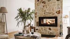 A cozy living room with a stone fireplace, a round wooden table with books and a candle, and a potted plant. - 'Serenity Gerringong' Designer Beach House (Werri Beach)