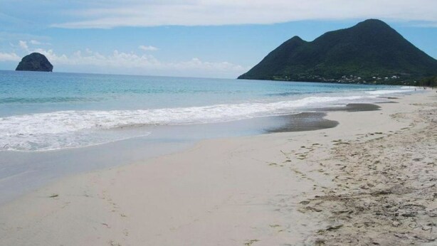 Une plage avec un ciel dégagé, une mer calme et deux montagnes imposantes en arrière-plan.