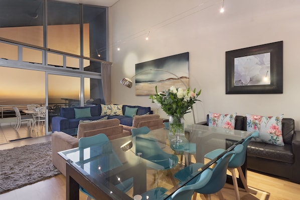 A modern living room with a glass dining table, blue chairs, a sofa with floral cushions, and a large window.