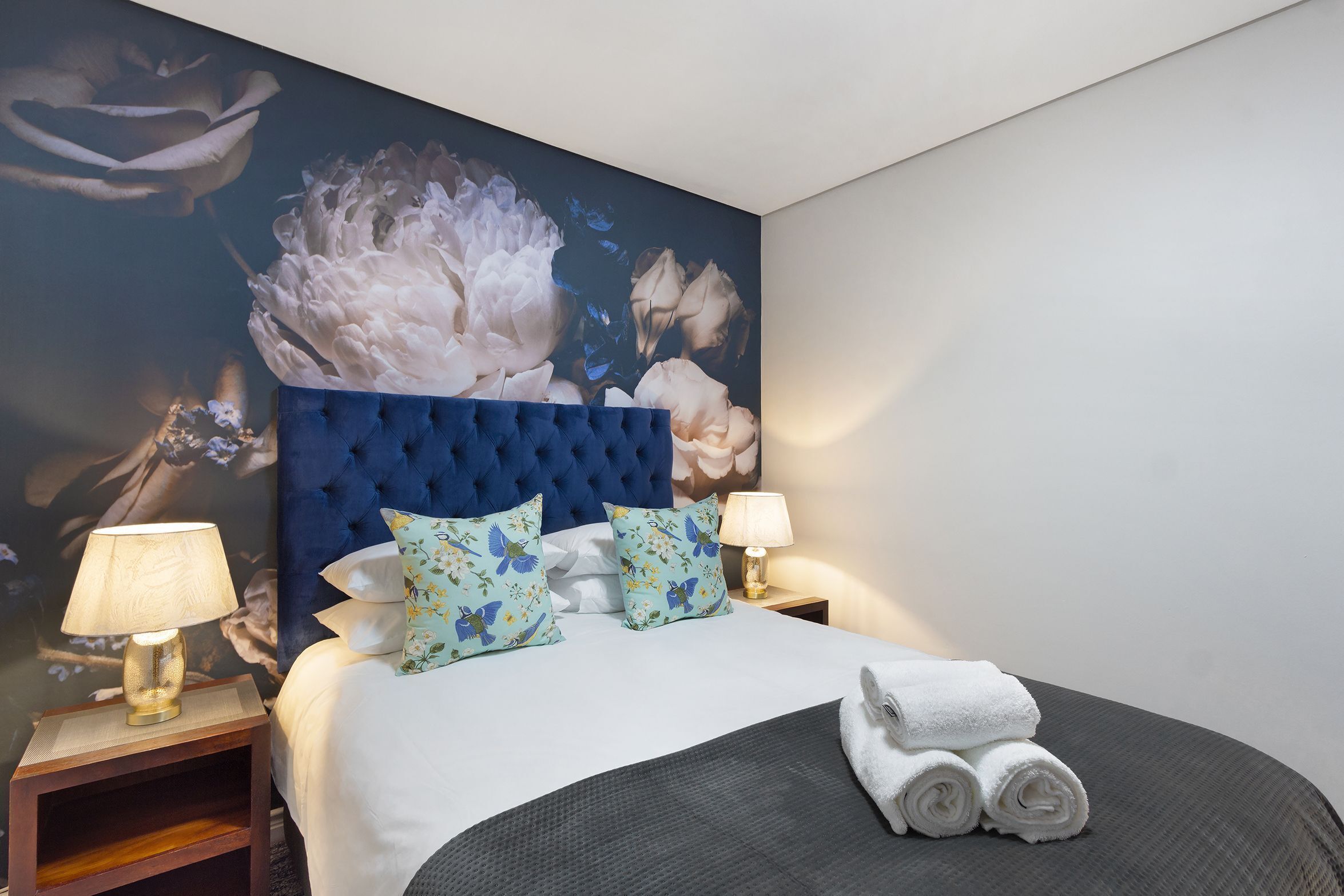 A hotel room with a large bed, two nightstands, a blue tufted headboard, floral wall art, and a pair of rolled towels on the bed.
