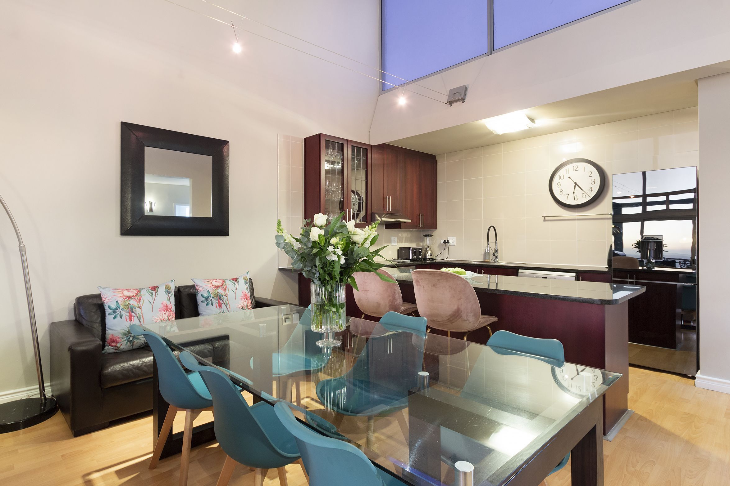 A modern kitchen with a glass dining table, blue chairs, and a vase of flowers.