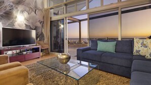 Una sala de estar moderna con un televisor grande, asientos cómodos, una mesa de centro de vidrio y vistas de la ciudad a través de amplios ventanales.