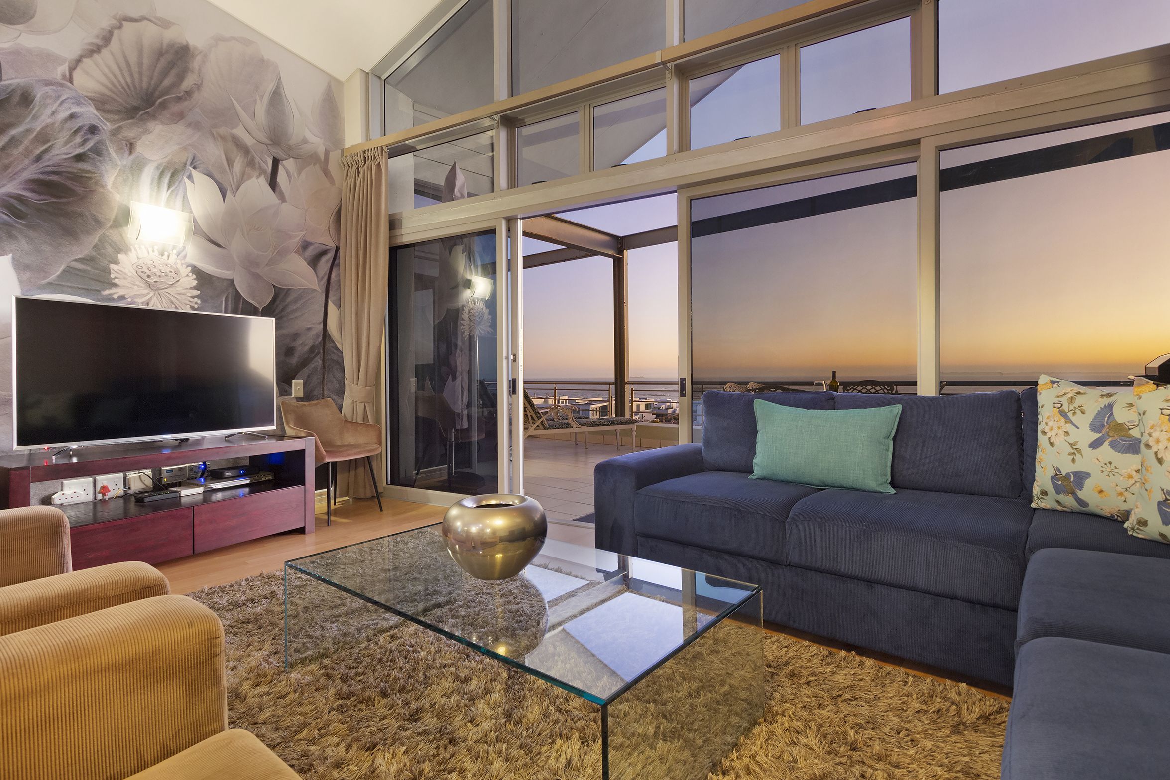 A modern living room with a large TV, comfortable seating, a glass coffee table, and a view of the cityscape through large windows.