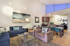 A modern living room with a sofa, armchairs, and a coffee table. There is a painting on the wall, a clock, and a kitchen area visible in the background.