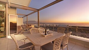 Balcón con una mesa lista para dos personas, con una botella y vasos, con vistas a una ciudad costera al atardecer.