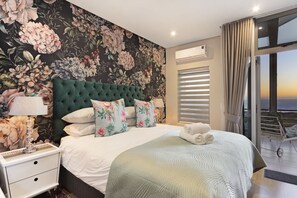 A bedroom with a large bed, floral wallpaper, a green headboard, and a view of the sea through the window. - Penthouse F23 Seaside Village (Cape Town)