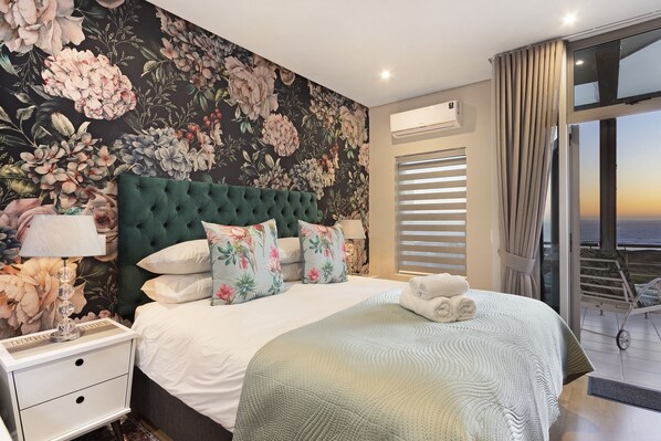 A bedroom with a large bed, floral wallpaper, a green headboard, and a view of the sea through the window.