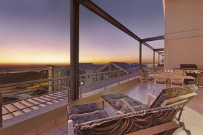 A balcony with outdoor seating, a view of the sunset, and a distant cityscape.