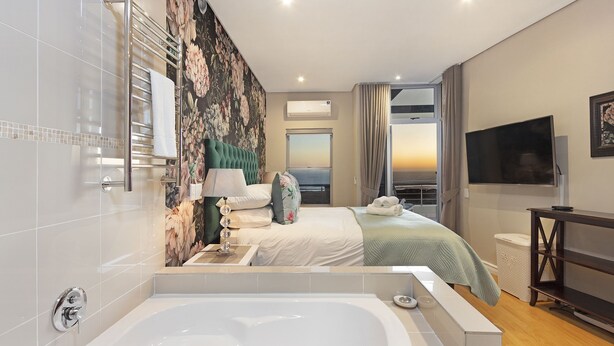Habitación de hotel moderna con una bañera grande, una cama con papel tapiz floral, un televisor y vistas de la puesta de sol a través de la ventana.