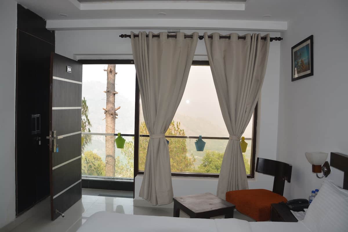 A hotel room with a bed, a desk, a chair, a window with curtains, a door, and a view of trees and mountains.