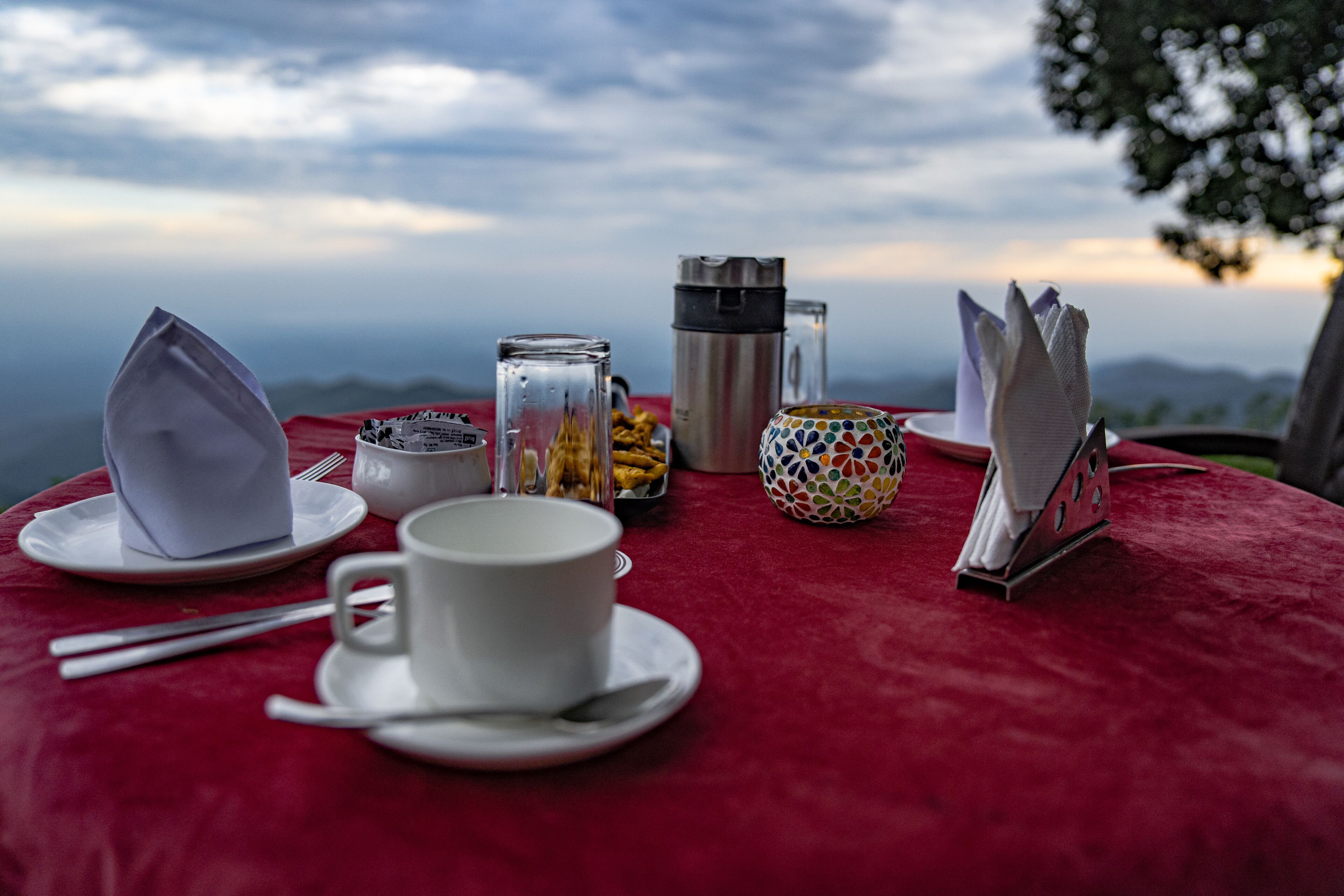 Una mesa dispuesta con una taza blanca, su platillo y una cuchara, una taza de colores, un termo, un frasco de vidrio, un recipiente metálico y un sujetavasos sobre un mantel rojo.