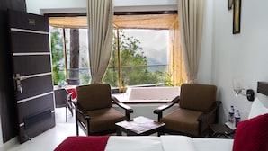Een hotelkamer met een bed, twee fauteuils, een tafeltje, een raam met gordijnen en uitzicht op bomen en bergen.