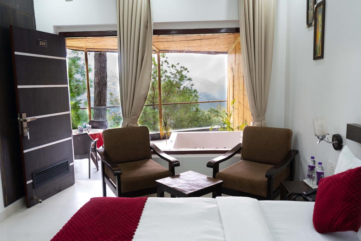 A hotel room with a bed, two armchairs, a small table, a window with curtains, and a view of trees and mountains.