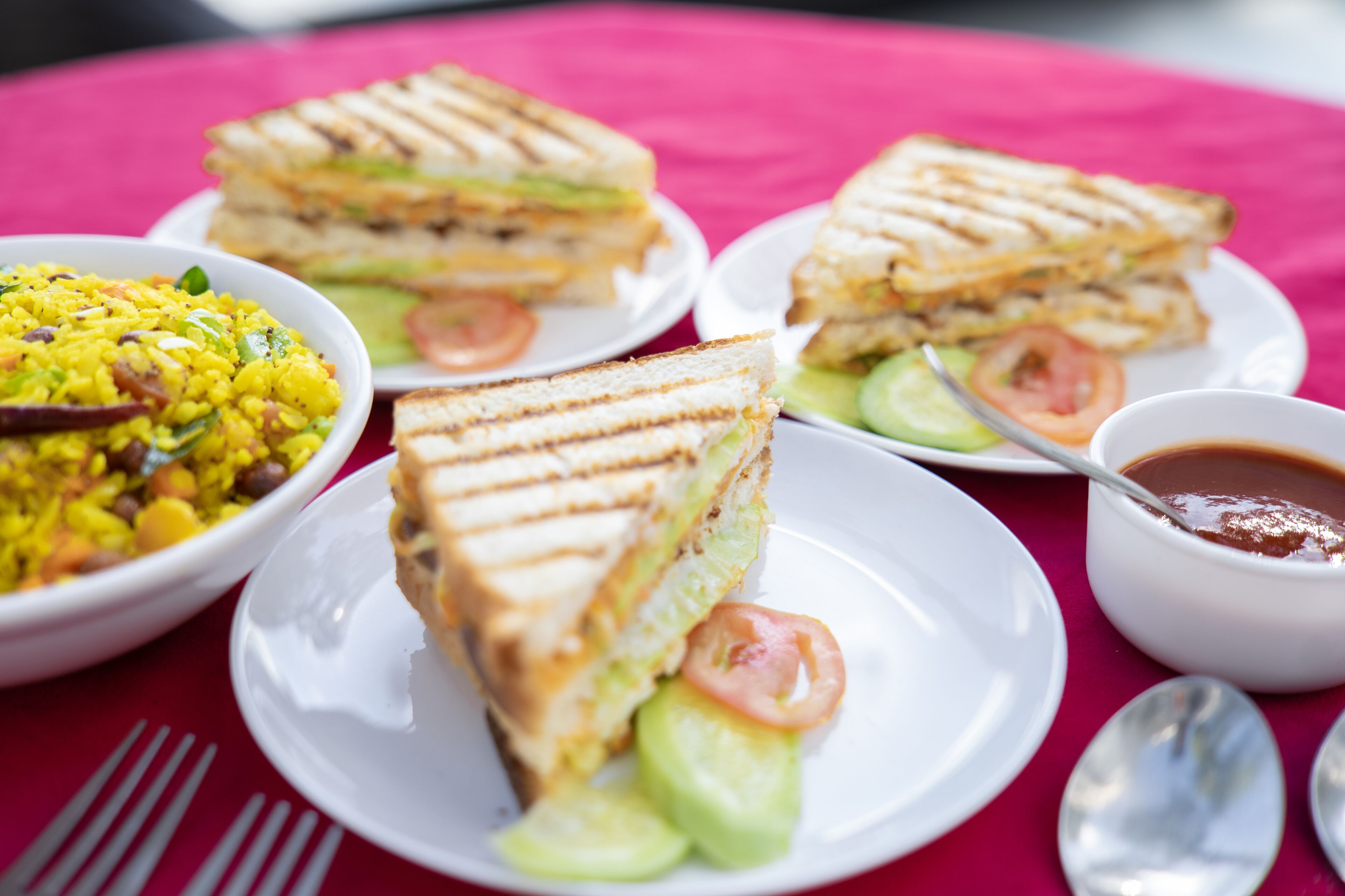 Una mesa con mantel rojo, un sándwich, un ensaladero, un plato con tomate y un recipiente pequeño con salsa.
