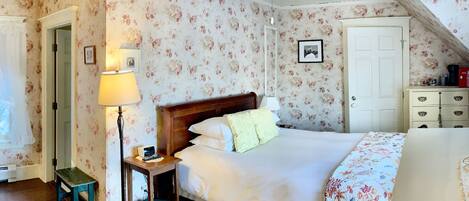 A bedroom with floral wallpaper, a wooden bed, a nightstand with a lamp, a ceiling fan, and a door with a picture frame.