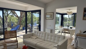 Ett modernt vardagsrum med en beige soffa, en trästol och ett soffbord i glas. I rummet finns även en matplats med ett bord och stolar, som syns genom stora glasdörrar.