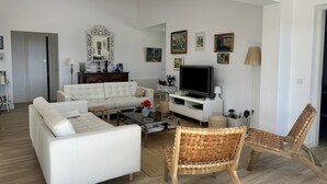 Ett rymligt vardagsrum med en vit soffa, en tv och ett soffbord. På väggen hänger inramade tavlor och ovanför soffan finns en spegel.