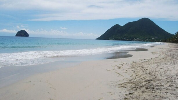 Una playa con arena, agua tranquila y una montaña al fondo.