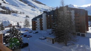 A snow-covered ski resort with modern buildings, a parking area, and a ski slope in the background.