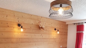 A cozy room with a wooden wall, a sofa with red and white patterned cushions, a dining table with chairs, and a pendant light fixture.