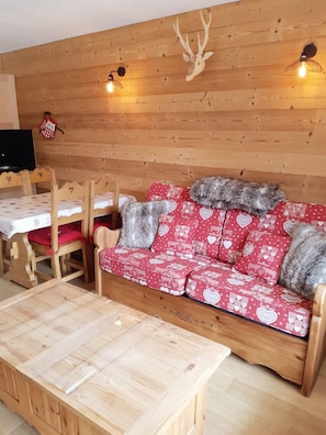 A cozy room with a wooden wall, a red patterned sofa with heart designs, a wooden coffee table, and a wooden dining table with chairs.