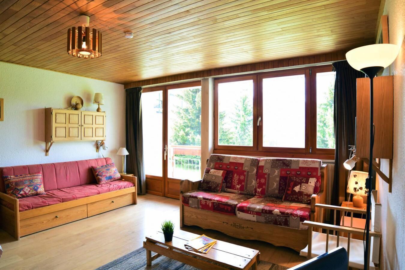 A cozy living room with a wooden ceiling, a red sofa, a patterned sofa, a wooden coffee table, and a window with a view of greenery.