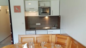 A compact kitchen with white cabinets, a built-in oven, and a microwave. There is a wooden table with chairs and a bench, and a blue couch in the foreground.