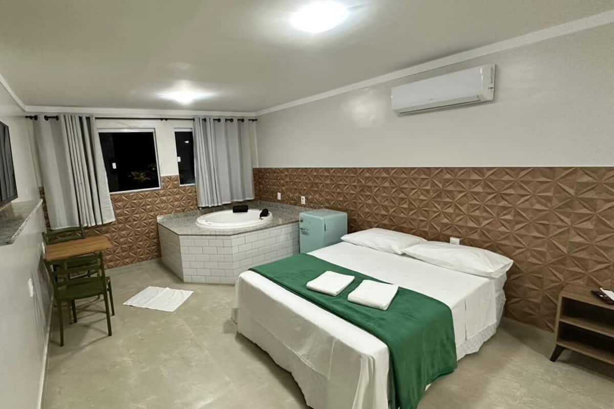 A hotel room with a bed, a desk, a chair, a window, a jacuzzi, and an air conditioning unit.