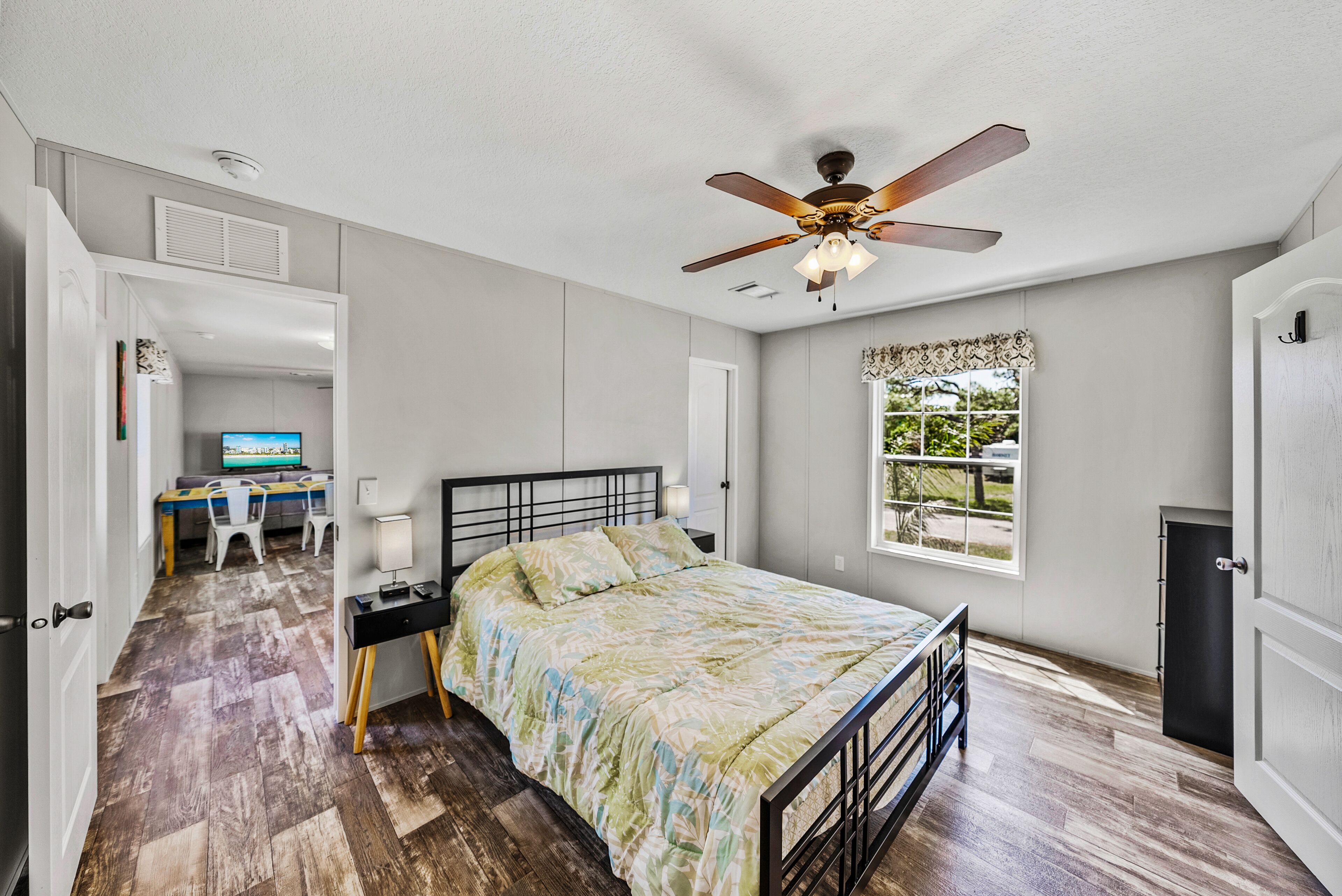 Un dormitorio con cama, ventilador de techo, ventana con vistas, escritorio con ordenador y puerta que da a otra habitación.
